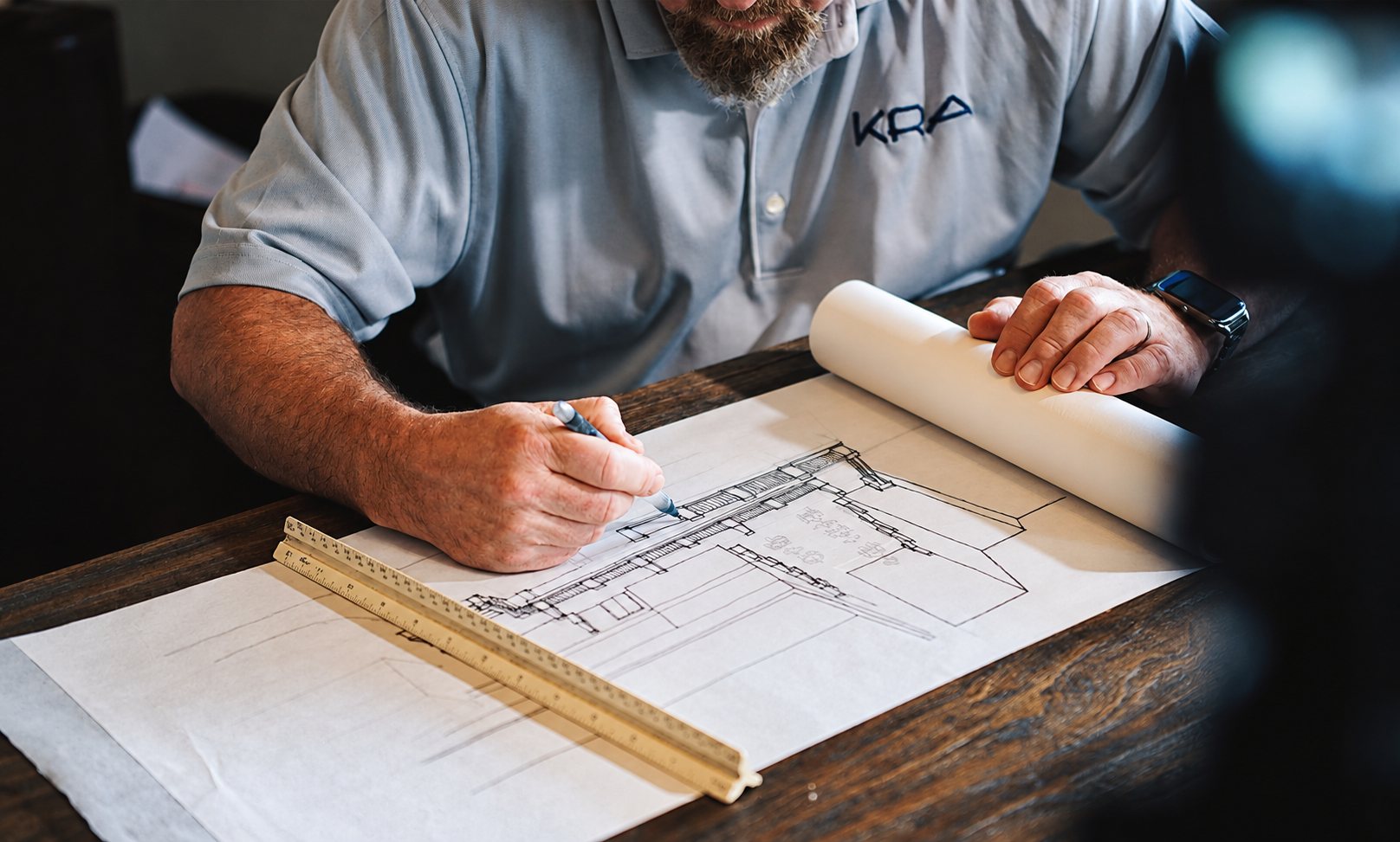Un homme en polo gris brodé KRA dessine au crayon un plan d'architecture sur une grande feuille posée sur une table en bois, avec une règle graduée et un rouleau de papier.
