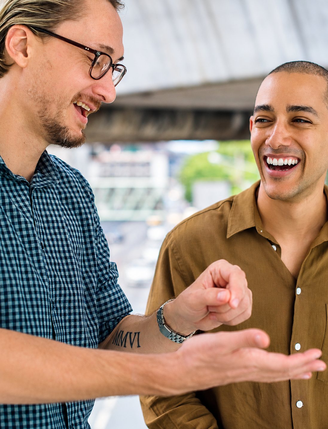 Deux hommes souriants discutent en riant à l'extérieur, l'un portant des lunettes et une chemise à carreaux, l'autre une chemise marron.